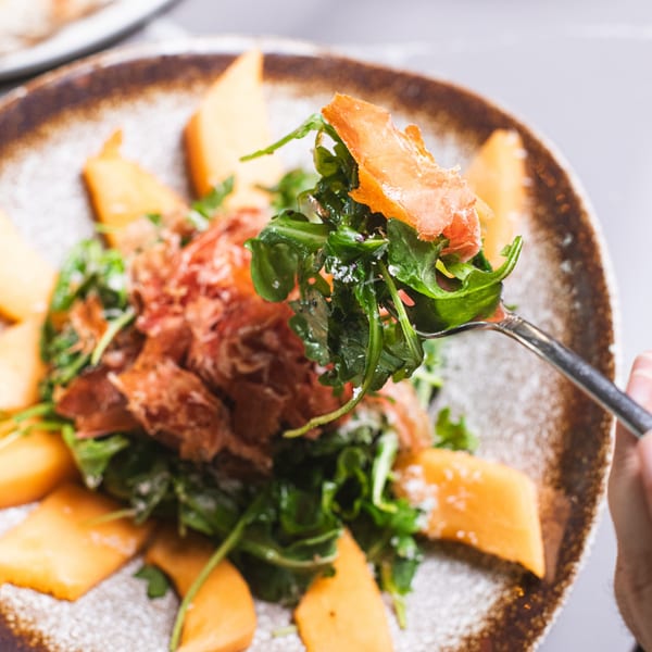 A fork lifts arugula and prosciutto from a da Tino. Italian salad with melon slices.