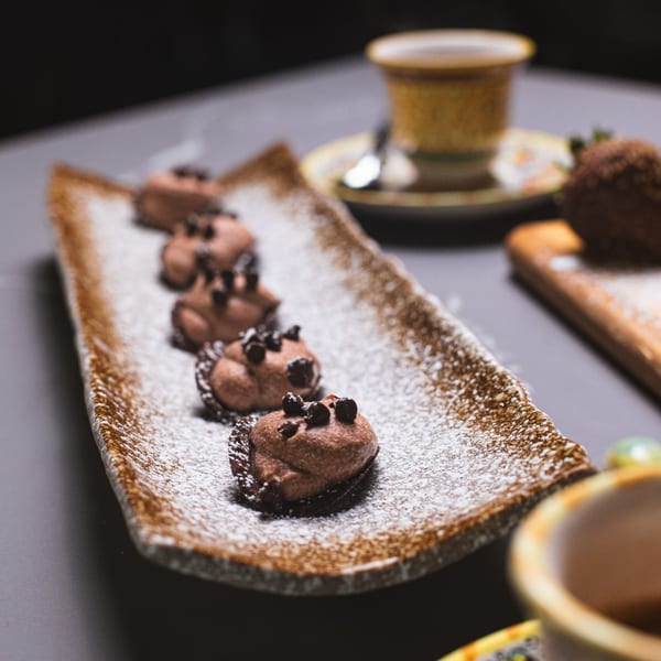 A long brown plate holds five chocolate desserts dusted with powdered sugar and chocolate chips.