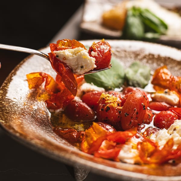 A spoon lifts roasted cherry tomatoes and creamy cheese from a rustic bowl, garnished with herbs.