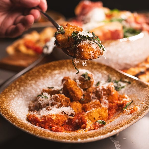 A spoon lifts a fried gnocchi from a bowl of gnocchi with red sauce, cheese, and basil.