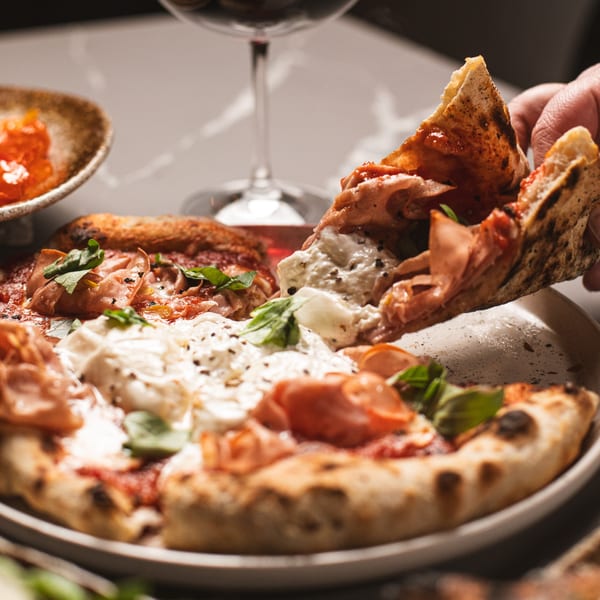 A hand lifts a slice of da Tino's Italian pizza with prosciutto, burrata, and basil.