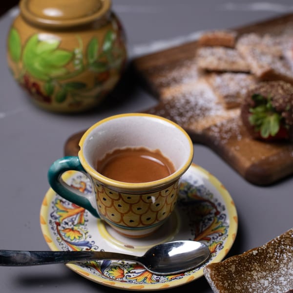 Espresso in a colorful ceramic cup with a spoon, alongside Italian pastries dusted with powdered sugar.