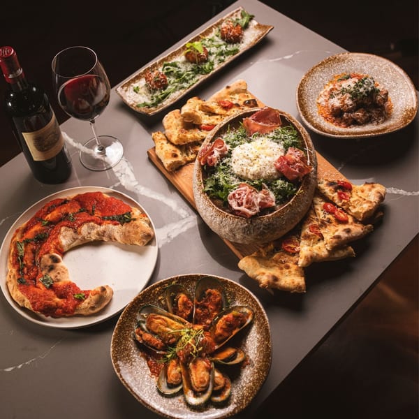 A table laden with various Italian dishes, including pizza, mussels, meatballs, and a salad, with a bottle of red wine.